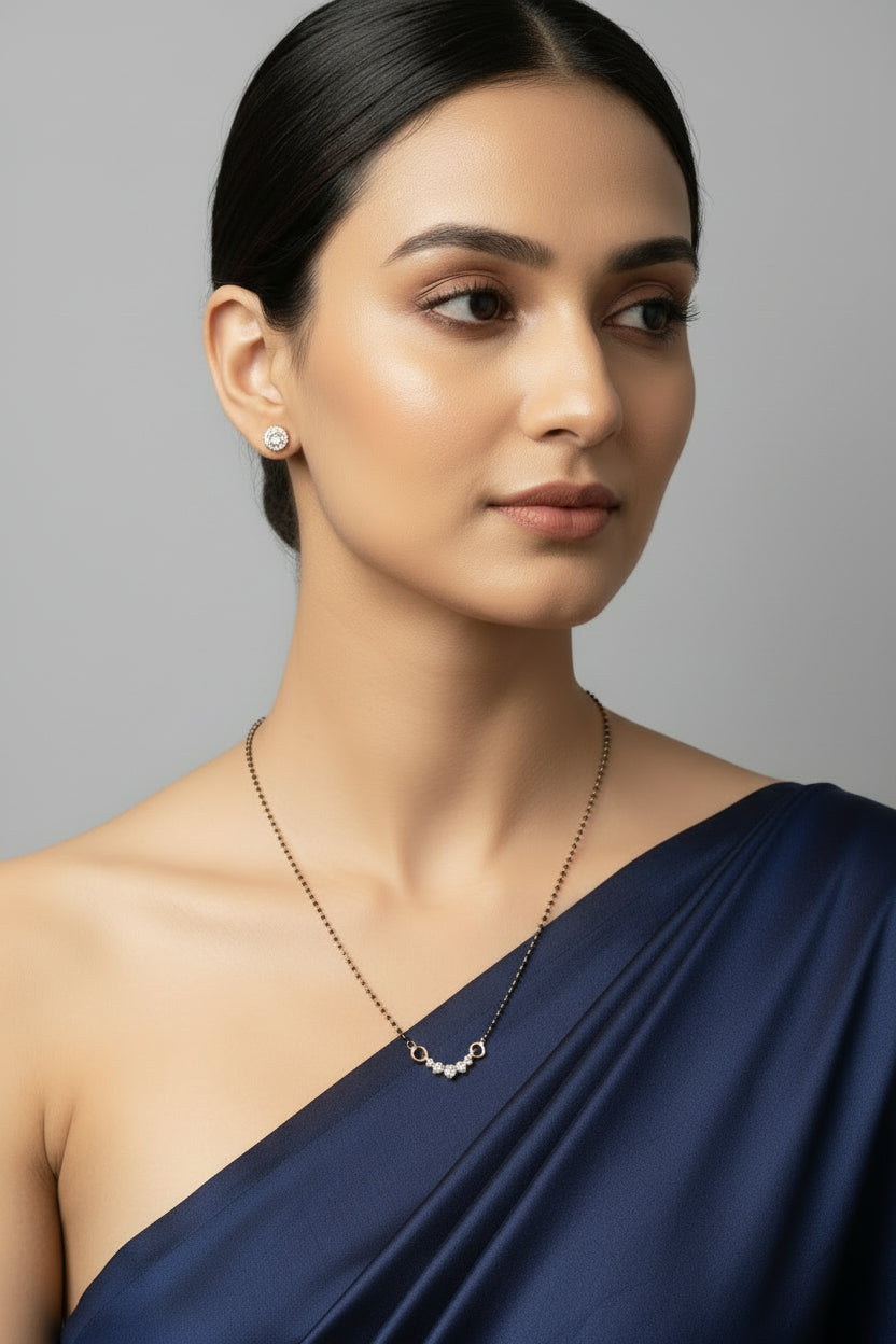 A delicate single-strand mangalsutra featuring a slim black bead chain and a central gold-toned arc set with five small, graduating clear crystals. The necklace is displayed on a black velvet jewelry bust against a dark background