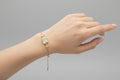 A delicate gold-toned adjustable chain bracelet featuring a central rectangular clear shimmering crystal. A small gold branch or leaf motif set with tiny clear stones is layered over the centerpiece, shown worn on a wrist against a soft purple background.