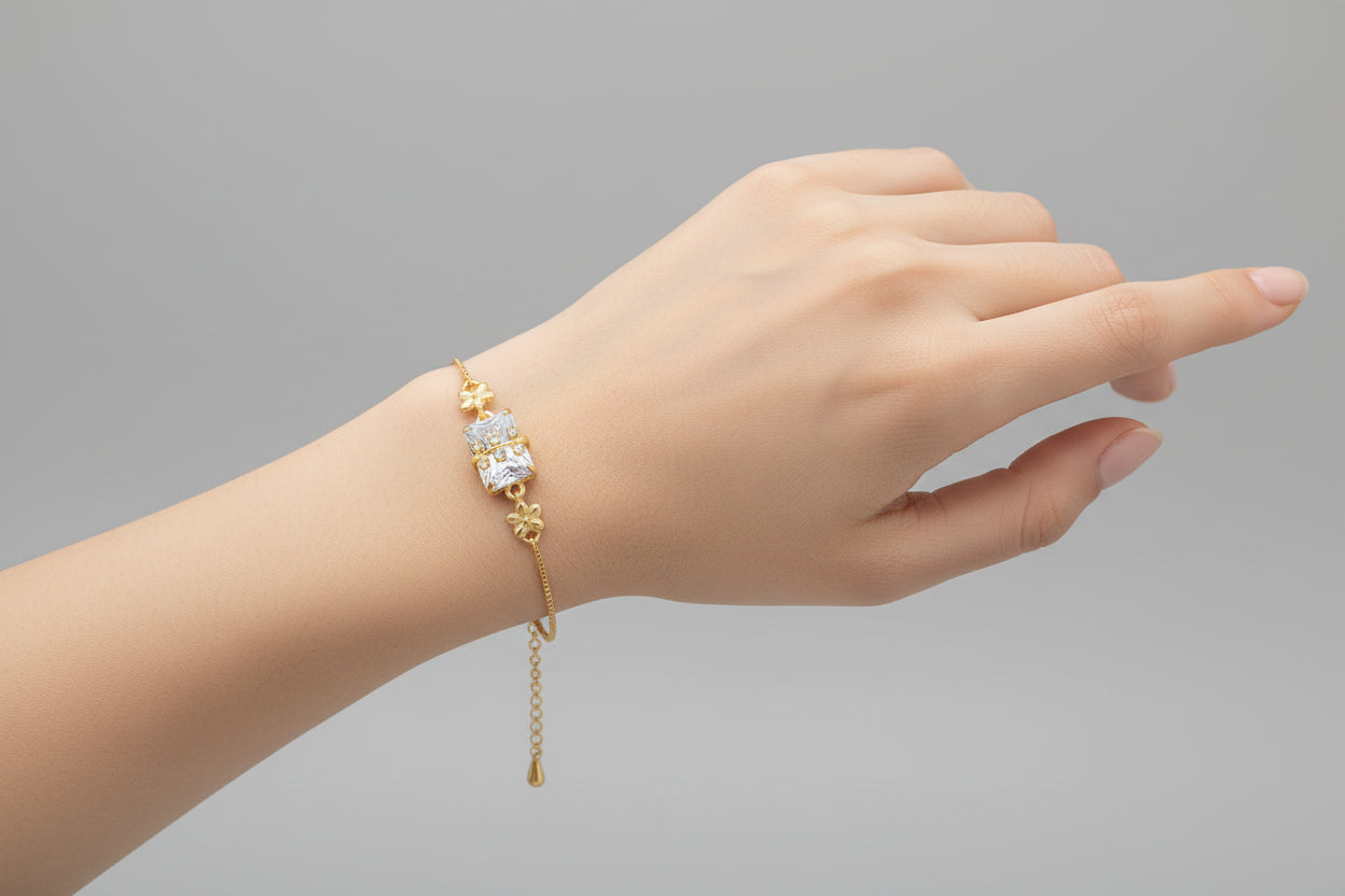 A delicate gold-toned adjustable chain bracelet featuring a central rectangular clear shimmering crystal. A small gold branch or leaf motif set with tiny clear stones is layered over the centerpiece, shown worn on a wrist against a soft purple background.