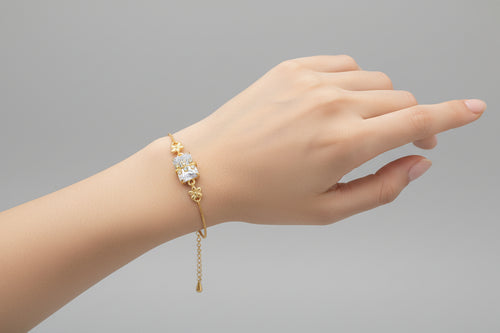 A delicate gold-toned adjustable chain bracelet featuring a central rectangular clear shimmering crystal. A small gold branch or leaf motif set with tiny clear stones is layered over the centerpiece, shown worn on a wrist against a soft purple background.