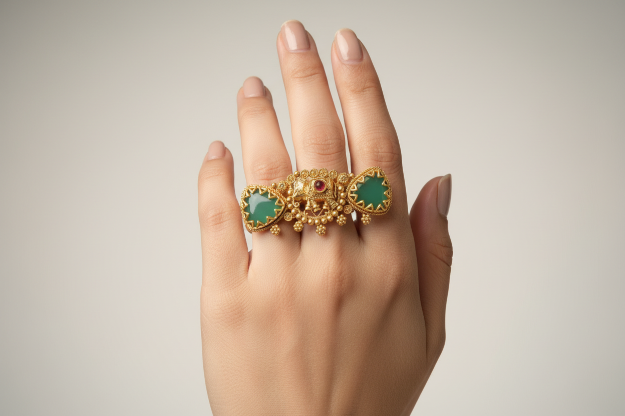 Antique gold-plated statement ring featuring a central elephant motif, two triangular green jade-style stones, and hanging gold bell charms, displayed against a natural leaf background.