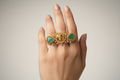 Antique gold-plated statement ring featuring a central elephant motif, two triangular green jade-style stones, and hanging gold bell charms, displayed against a natural leaf background.