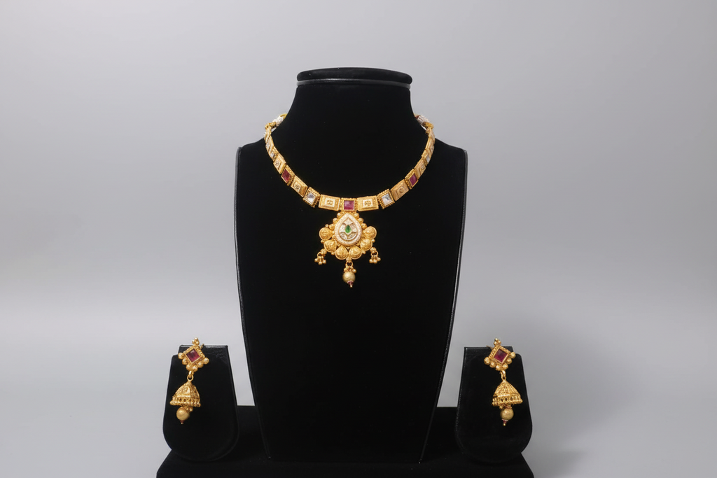 A traditional gold-toned jewelry set featuring a necklace with square stations set with ruby-red and clear stones, leading to a large, intricate gold pendant. The set includes matching Jhumka earrings with red stone accents, displayed on a black jewelry bust.
