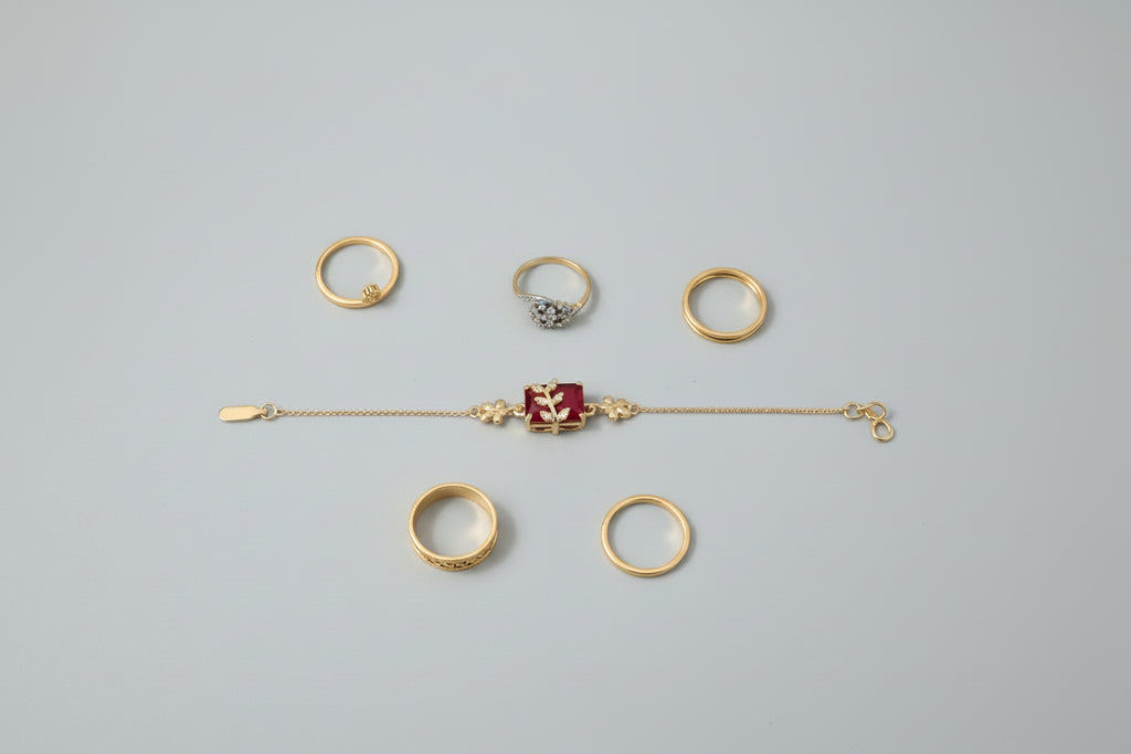A dainty gold-toned chain bracelet featuring a central rectangular ruby-red stone. A small, gold leaf-shaped motif encrusted with tiny clear crystals is set atop the red stone, worn on a wrist against a vibrant purple background.