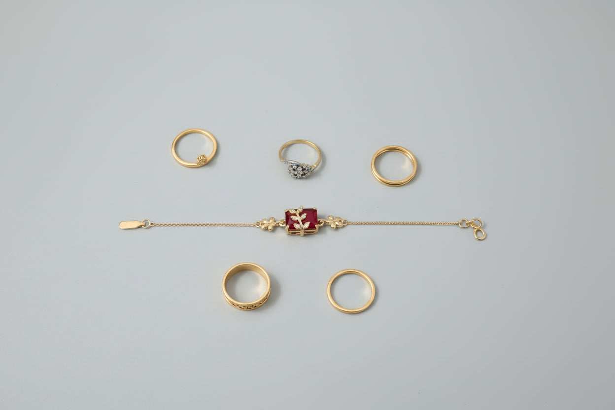 A dainty gold-toned chain bracelet featuring a central rectangular ruby-red stone. A small, gold leaf-shaped motif encrusted with tiny clear crystals is set atop the red stone, worn on a wrist against a vibrant purple background.
