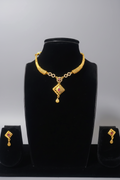 A gold-toned jewelry set featuring a necklace with a textured tubular collar and a diamond-shaped pendant. The pendant has a central ruby-red stone and intricate cutout detailing. The set includes matching diamond-shaped drop earrings, displayed on a black jewelry bust.