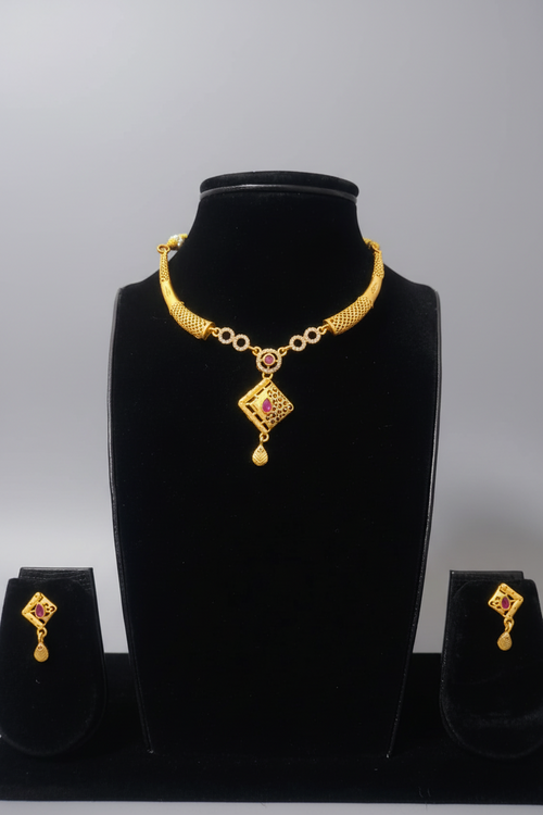 A gold-toned jewelry set featuring a necklace with a textured tubular collar and a diamond-shaped pendant. The pendant has a central ruby-red stone and intricate cutout detailing. The set includes matching diamond-shaped drop earrings, displayed on a black jewelry bust.