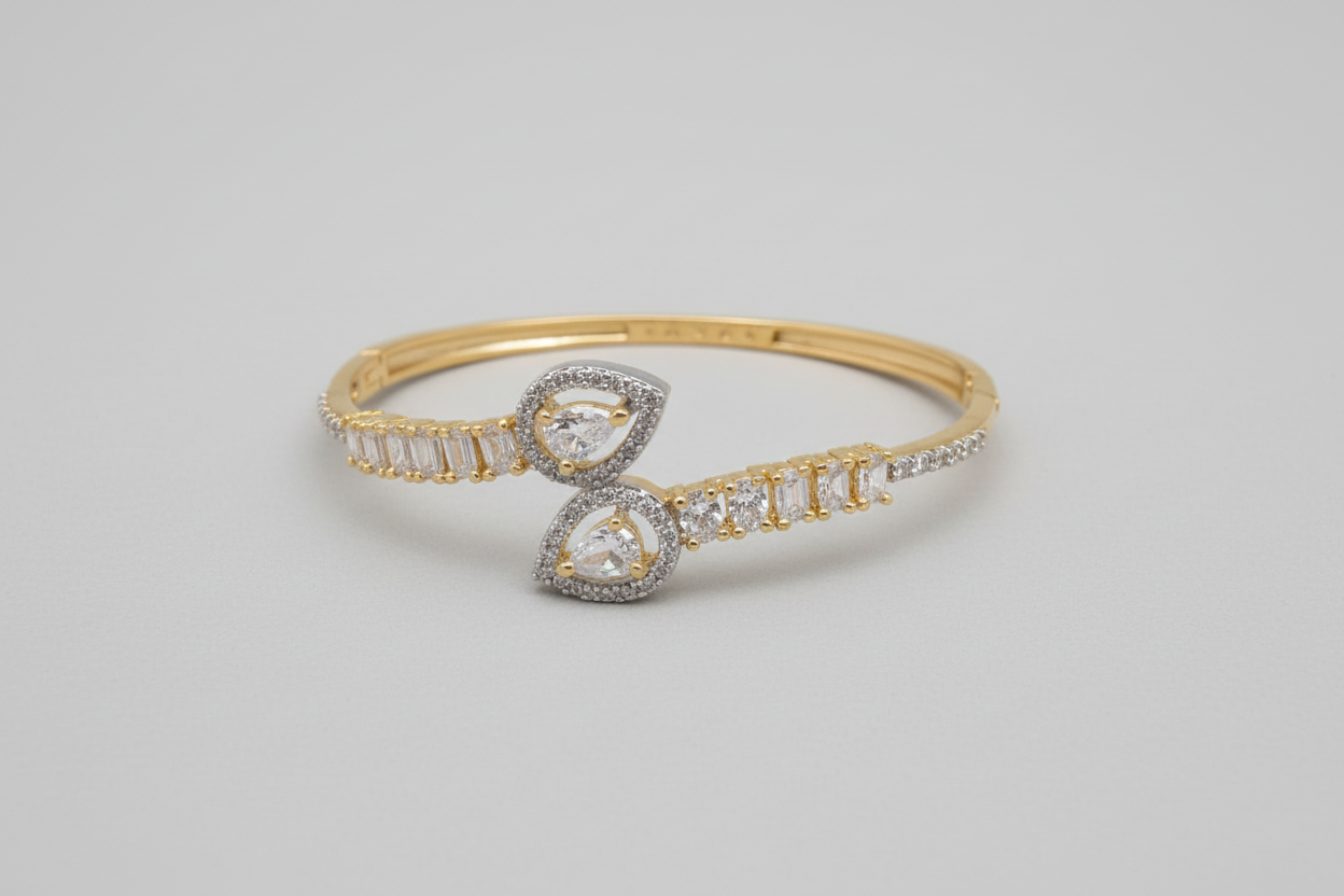A gold-toned bypass-style bangle featuring a split band encrusted with a row of clear crystals, terminating in two large teardrop-shaped motifs also set with shimmering stones, displayed on a dark green velvet surface.