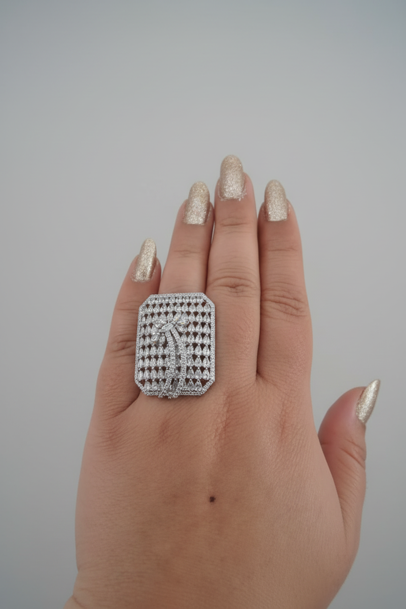 A large, oversized rectangular statement ring in a silver-tone finish, densely pavé-set with clear shimmering crystals. The center features a stylized palm tree or feather motif, shown worn on a hand with gold-toned nails against a dark background
