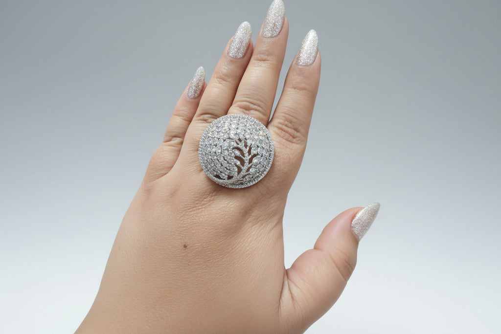 A large, oversized circular statement ring in a silver-tone finish, densely pavé-set with clear shimmering crystals. The design features an intricate tree-of-life or branch motif within the circular frame, shown worn on a hand against a dark background