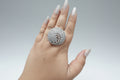 A large, oversized circular statement ring in a silver-tone finish, densely pavé-set with clear shimmering crystals. The design features an intricate tree-of-life or branch motif within the circular frame, shown worn on a hand against a dark background
