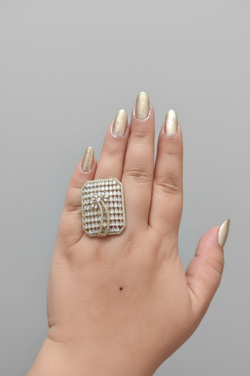 A large, oversized rectangular statement ring in a silver-tone finish, densely pavé-set with clear shimmering crystals. The center features a stylized palm tree or feather motif, shown worn on a hand with gold-toned nails against a dark background.