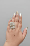 A large, oversized rectangular statement ring in a silver-tone finish, densely pavé-set with clear shimmering crystals. The center features a stylized palm tree or feather motif, shown worn on a hand with gold-toned nails against a dark background.