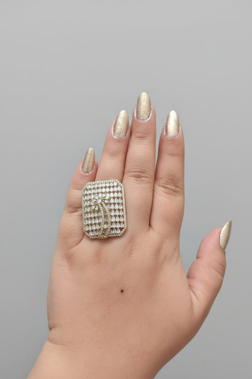 A large, oversized rectangular statement ring in a silver-tone finish, densely pavé-set with clear shimmering crystals. The center features a stylized palm tree or feather motif, shown worn on a hand with gold-toned nails against a dark background.