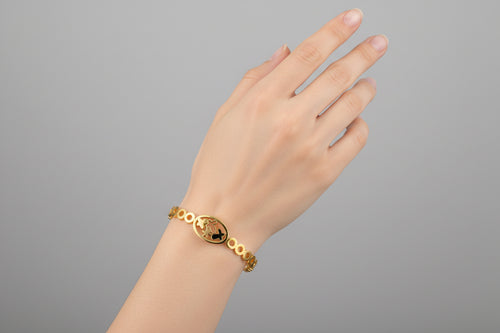 A dainty gold-toned open-cuff bangle featuring a central multi-petaled floral motif and two smaller four-leaf clover charms on either side, displayed on a vibrant pink background