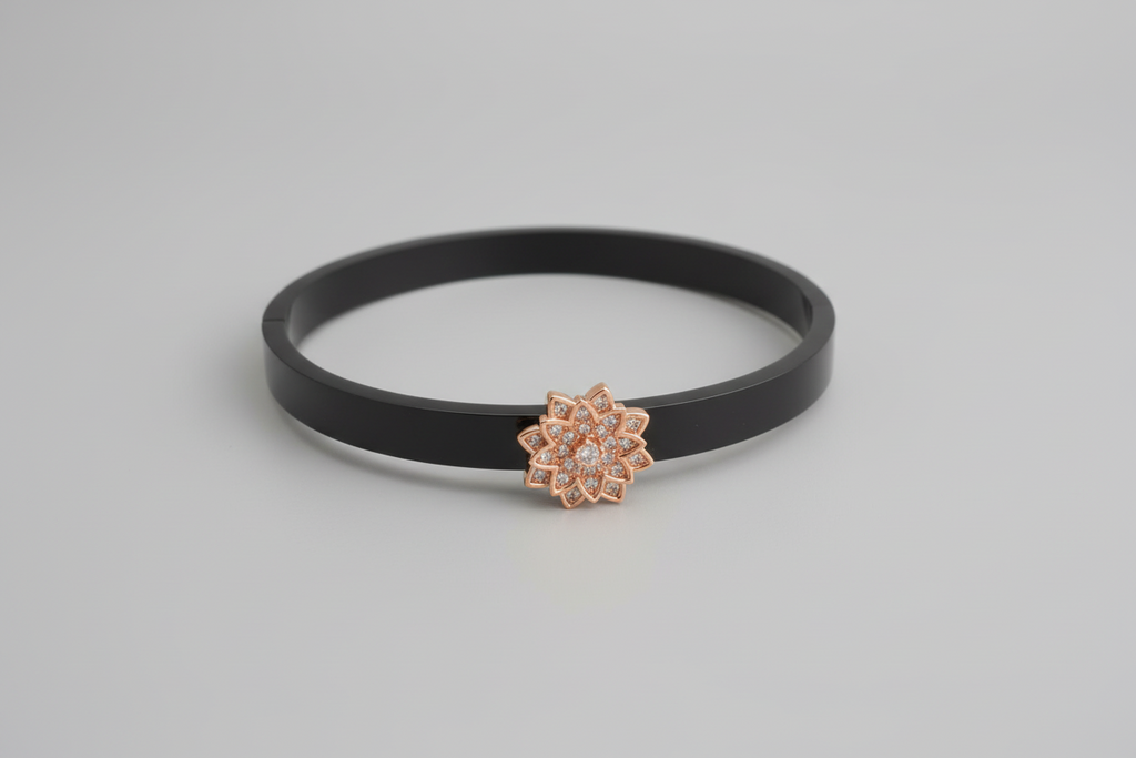 A wide, sleek black bangle featuring a central rose-gold floral motif encrusted with small crystals, resting on a dark green textured surface.