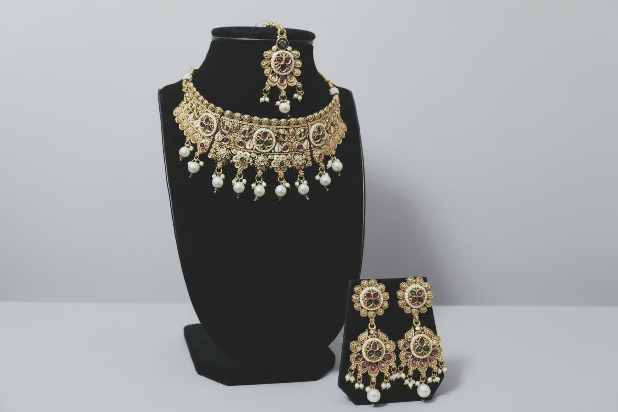 A heavy, traditional antique gold-finished choker necklace and matching maang tikka set. The design features intricate floral and circular metalwork, accented with green, pink, and clear stones, and finished with multiple white pearl droplets, displayed on a black jewelry bust.