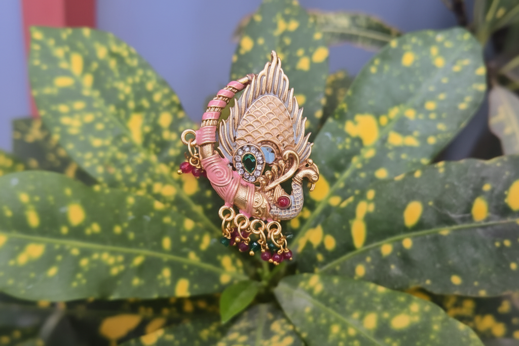 Large gold-plated statement ring featuring a peacock motif with pink enamel work, a green center stone, and dangling pink and green beads, photographed on a vibrant green leaf.