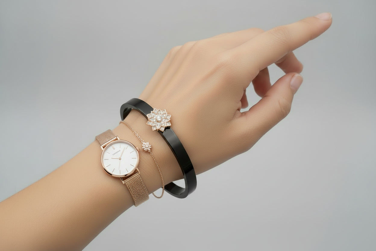 A wide, sleek black bangle featuring a central rose-gold floral motif encrusted with small crystals, resting on a dark green textured surface.