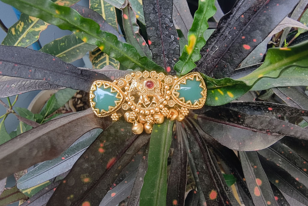 Antique gold-plated statement ring featuring a central elephant motif, two triangular green jade-style stones, and hanging gold bell charms, displayed against a natural leaf background.