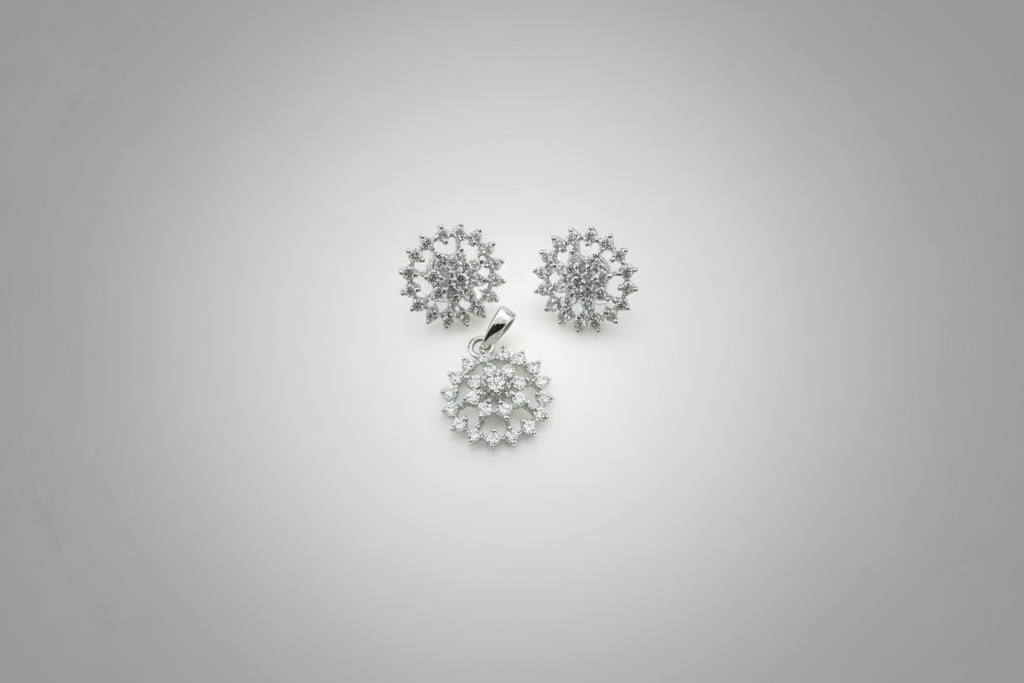 A three-piece jewelry set in a silver-tone finish, featuring a circular pendant and two matching stud earrings. Each piece is intricately set with clear shimmering crystals in a floral or snowflake-like pattern, displayed against a dark black background.