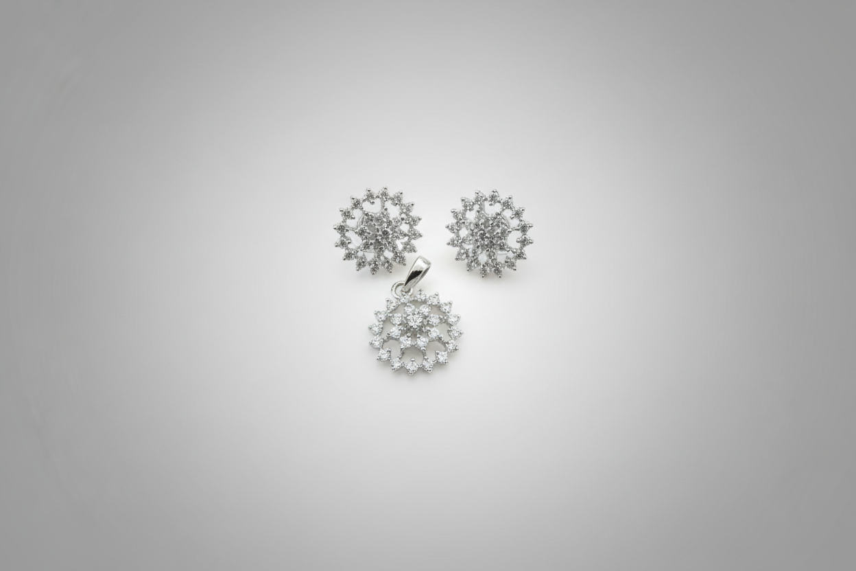 A three-piece jewelry set in a silver-tone finish, featuring a circular pendant and two matching stud earrings. Each piece is intricately set with clear shimmering crystals in a floral or snowflake-like pattern, displayed against a dark black background.