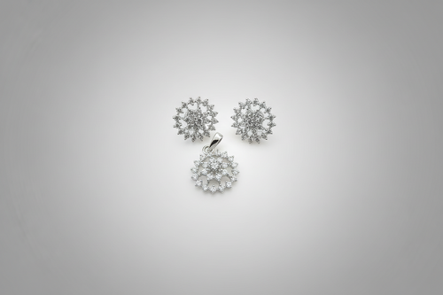 A three-piece jewelry set in a silver-tone finish, featuring a circular pendant and two matching stud earrings. Each piece is intricately set with clear shimmering crystals in a floral or snowflake-like pattern, displayed against a dark black background.