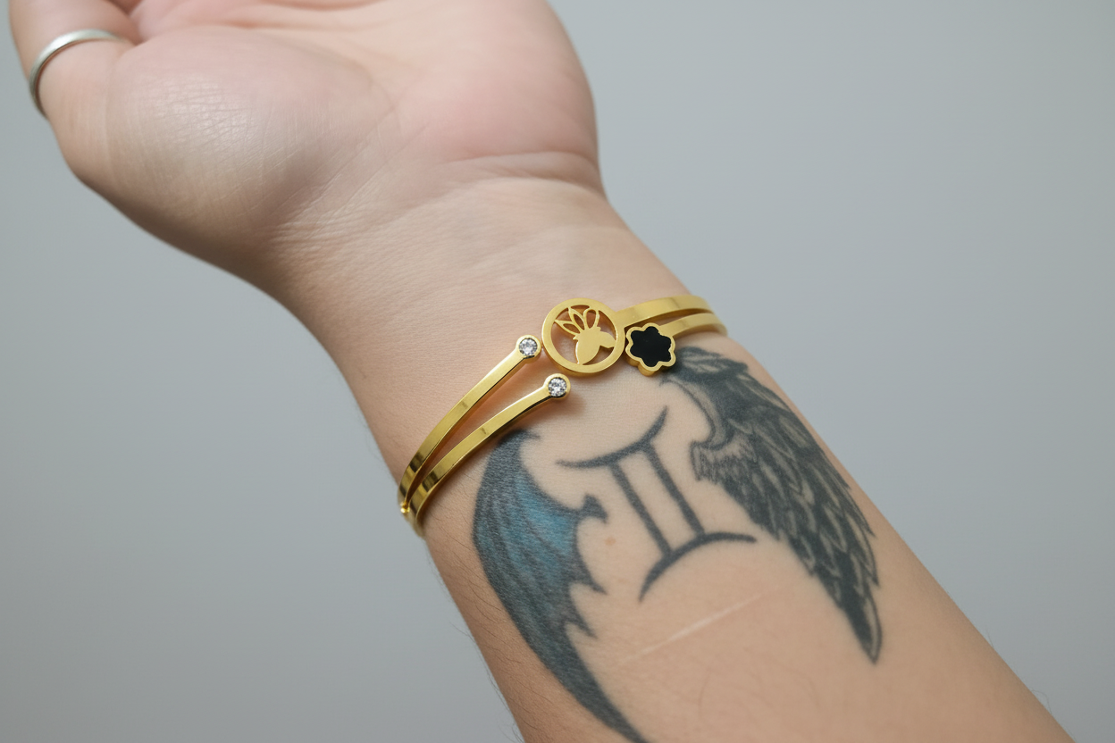 A delicate gold-toned bangle featuring a split-band design. The centerpiece includes a black four-leaf clover charm and a circular cutout charm with a crescent moon and star motif, shown worn on a wrist against a vibrant pink background
