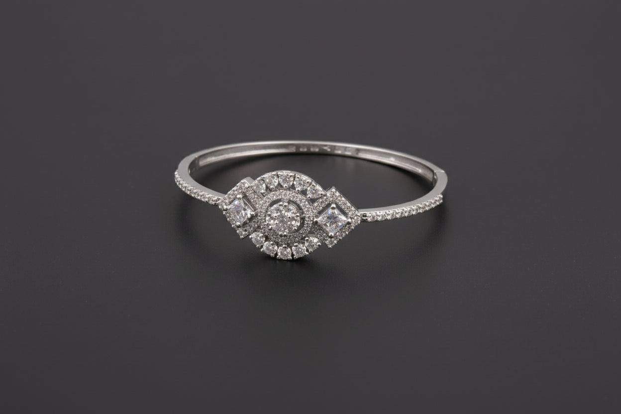 A silver-toned statement bangle with a large central circular cluster of clear crystals flanked by two diamond-shaped stones, featuring a fully encrusted band on a dark textured surface