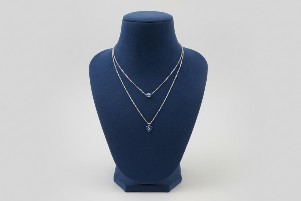 A silver-toned double-layered necklace featuring two distinct blue evil eye pendants. The upper shorter chain holds a round blue nazar bead, while the longer lower chain features a dark blue heart-shaped evil eye pendant. Displayed on a navy blue jewelry mannequin