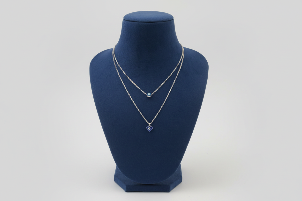 A silver-toned double-layered necklace featuring two distinct blue evil eye pendants. The upper shorter chain holds a round blue nazar bead, while the longer lower chain features a dark blue heart-shaped evil eye pendant. Displayed on a navy blue jewelry mannequin
