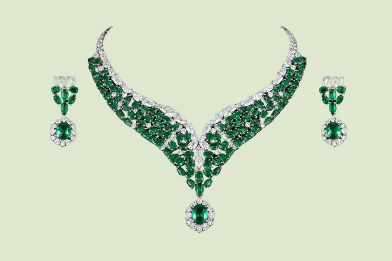 A luxury jewelry set featuring a V-shaped necklace and matching drop earrings, both adorned with vibrant green emerald-cut gemstones and brilliant clear stones in a silver-toned setting, presented on a solid gray background.