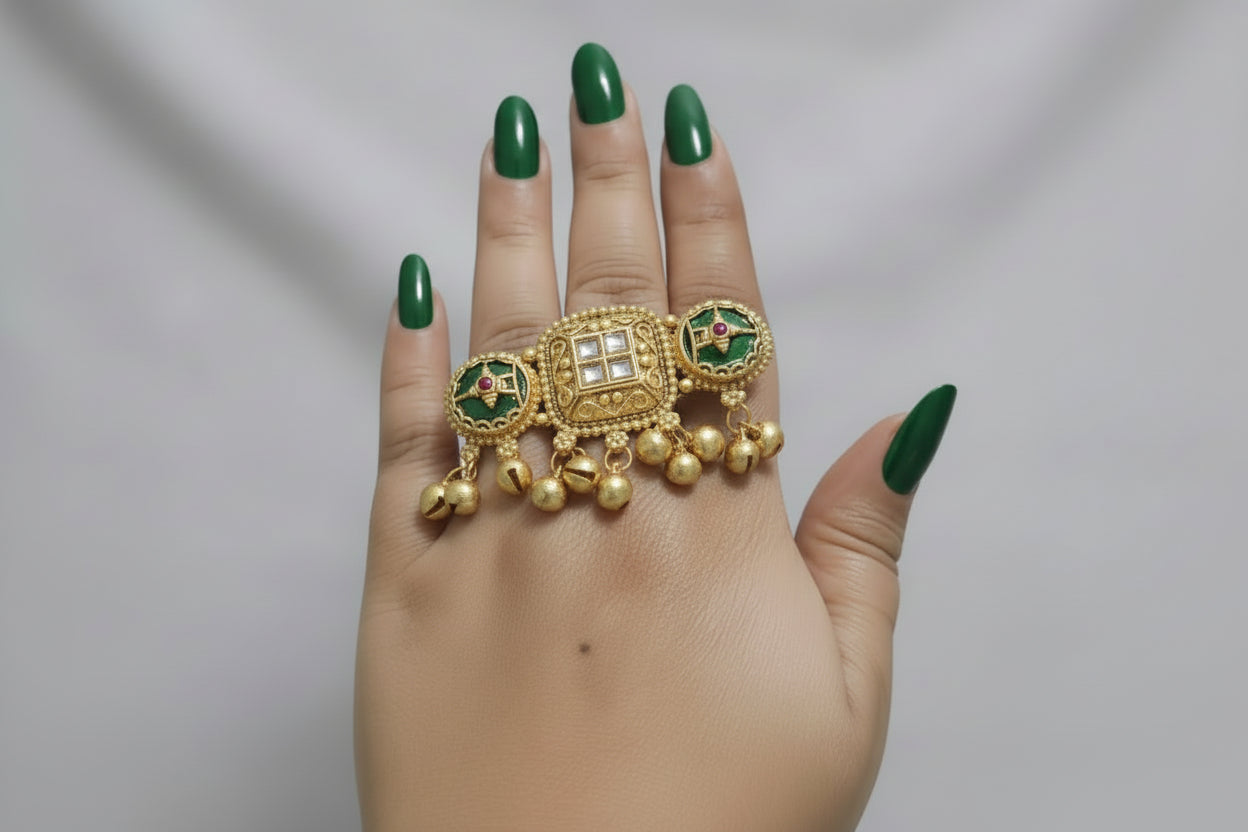 A wide, horizontal two-finger statement ring in antique gold finish, featuring a central square Kundan-style stone, green enamel side circles with pink accents, and multiple hanging gold ghungroo beads, worn on a hand with gold glitter nails.