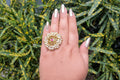 A large, circular gold-toned statement ring featuring a dense cluster of small white seed pearls surrounding a central ruby-red stone. The ring is worn on a hand with gold-glitter nails, posed against a backdrop of green and yellow speckled leaves