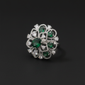 A large, oversized circular statement ring in a silver-tone finish, featuring an intricate openwork floral design set with clear crystals and multiple teardrop and oval-cut emerald-green stones. The ring is shown worn on a hand against a dark green velvet background