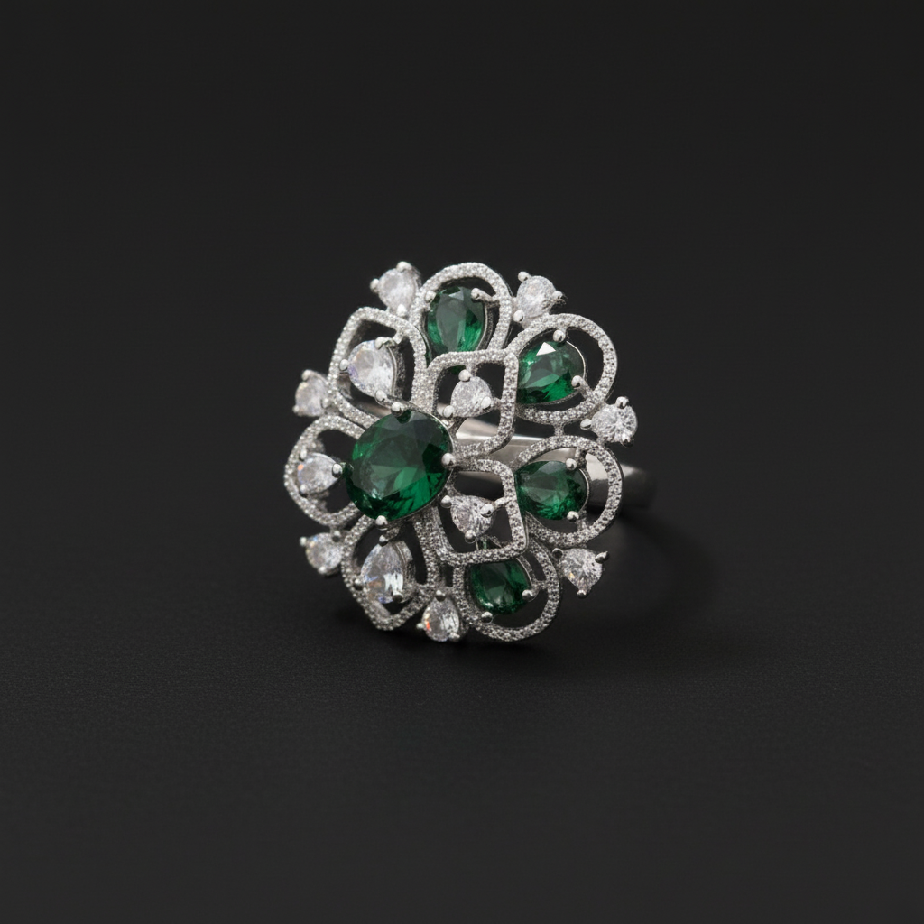 A large, oversized circular statement ring in a silver-tone finish, featuring an intricate openwork floral design set with clear crystals and multiple teardrop and oval-cut emerald-green stones. The ring is shown worn on a hand against a dark green velvet background