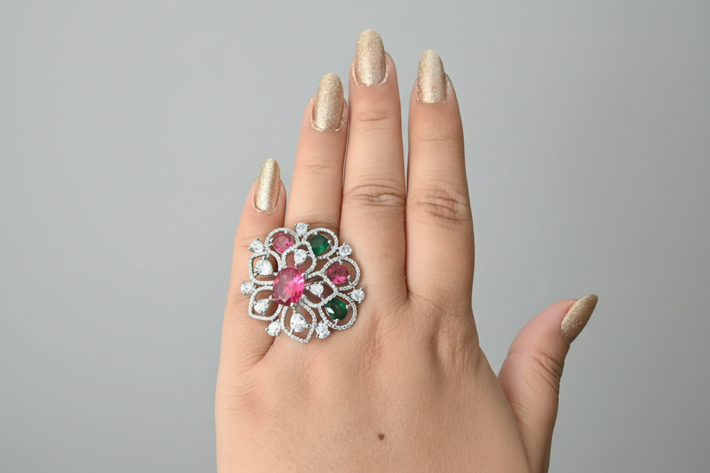 A large, circular silver-toned statement ring featuring an intricate floral lattice design. The ring is set with clear shimmering crystals and accented with oval-cut ruby-red and emerald-green stones, shown worn on a hand against a dark background.