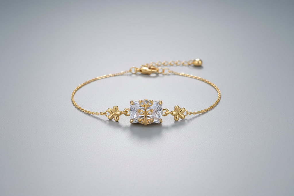 A delicate gold-toned adjustable chain bracelet featuring a central rectangular clear shimmering crystal. A small gold branch or leaf motif set with tiny clear stones is layered over the centerpiece, shown worn on a wrist against a soft purple background.