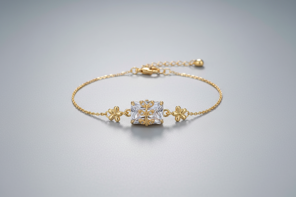A delicate gold-toned adjustable chain bracelet featuring a central rectangular clear shimmering crystal. A small gold branch or leaf motif set with tiny clear stones is layered over the centerpiece, shown worn on a wrist against a soft purple background.